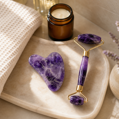 Purple amethyst facial roller and matching heart-shaped stone tool arranged on a beige tray beside a small amber candle jar, with a cream textured cloth and soft floral decor in the background.