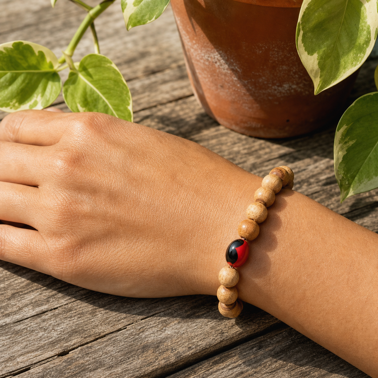 A wooden bead bracelet with a red and black ladybug charm worn on a person's wrist, resting on a weathered wooden surface with a potted plant in the background.