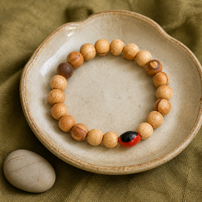 A wooden bead bracelet with light tan round beads and one red-and-black accent bead, displayed on a ceramic dish over an olive fabric background.