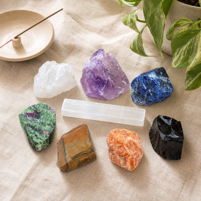 A set of seven colorful raw crystals and a white quartz stick placed on a beige linen cloth, with a small wooden dish and green plant leaves in the background.