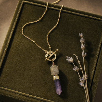A gold chain necklace with a toggle clasp and a pendant featuring a clear stone and a purple amethyst point, displayed inside a dark green velvet jewelry box next to dried lavender.