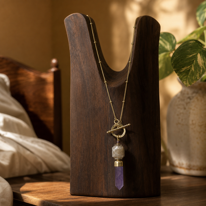 A gold-tone toggle necklace with a crescent moon and purple crystal pendant, displayed on a dark wooden stand in a warm, softly lit bedroom setting.