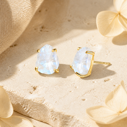 A pair of gold stud earrings with iridescent light-blue cabochon gemstones, styled on a textured beige stone surface with soft cream flowers and draped fabric.