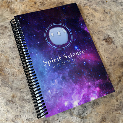 A purple-colored dotted journal with a spiral binding and 'Spirit Science Journal' printed on the cover, featuring a cosmic or space-themed design.